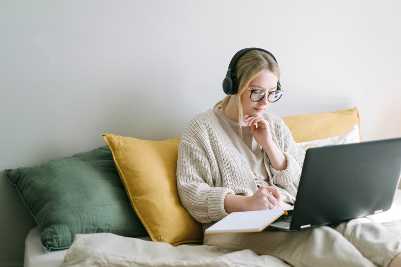 A person sits on a sofa with headphones, a notebook, and a computer to answer creative writing prompts