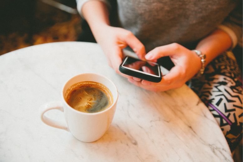 a woman holds a mobile phone answering shadow work journaling prompts with a cup of coffee nearby